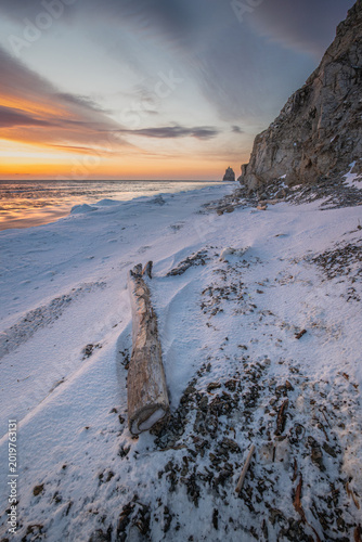 Snow-covered beach at sunset with a large driftwood log on the shore and rocky cliffs in the background, creating a serene coastal landscape