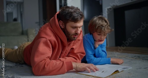 Father and young son lying on a living room rug, reading an open book together during a quiet family learning moment at home