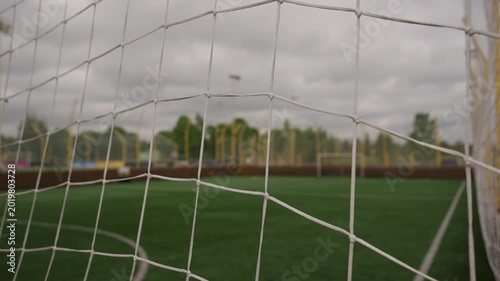 Small empty football training pitch with green turf and goals, surrounded by a white net fence. Strong wind moves the net, with a large stadium and floodlights visible in the background.