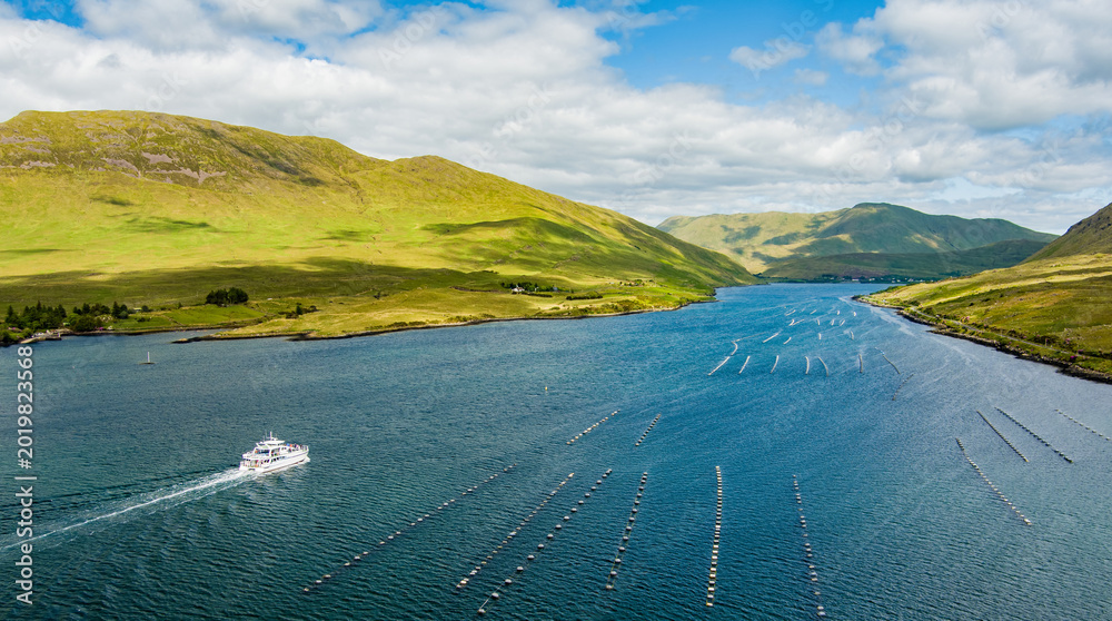 Fototapeta premium A tour boat cruises along Killary Fjord beneath bright green mountains and open skies in western Ireland. A scenic travel landscape highlighting sightseeing on Irelands only fjord.