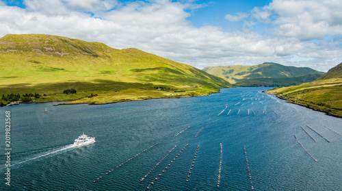 A tour boat cruises along Killary Fjord beneath bright green mountains and open skies in western Ireland. A scenic travel landscape highlighting sightseeing on Irelands only fjord.