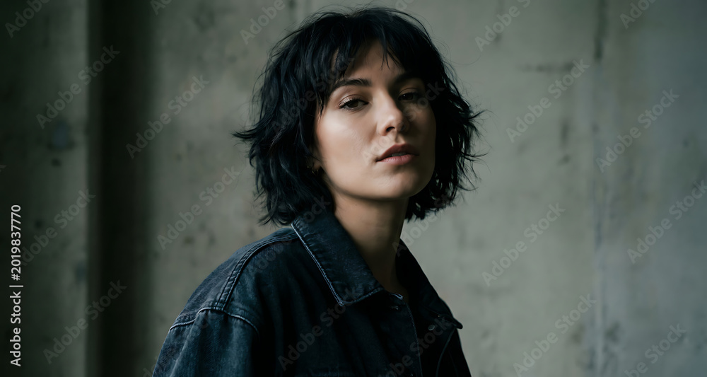 Fototapeta premium Portrait of Young Woman with Dark Hair and Denim Jacket Against Concrete Wall