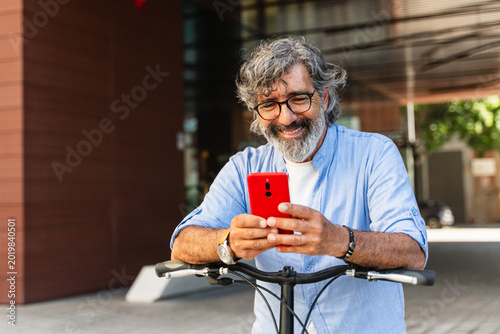 Photos Happy senior man with bicycle smiling while checking smartphone app on a modern