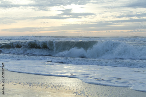 Foto Ocean waves crashing on a sandy beach at sunrise with soft clouds in the sky