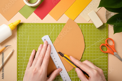 Hands using utility knife and ruler to cut colored paper on green cutting mat. Creative workspace with scissors, glue, and stationery for DIY craft projects.