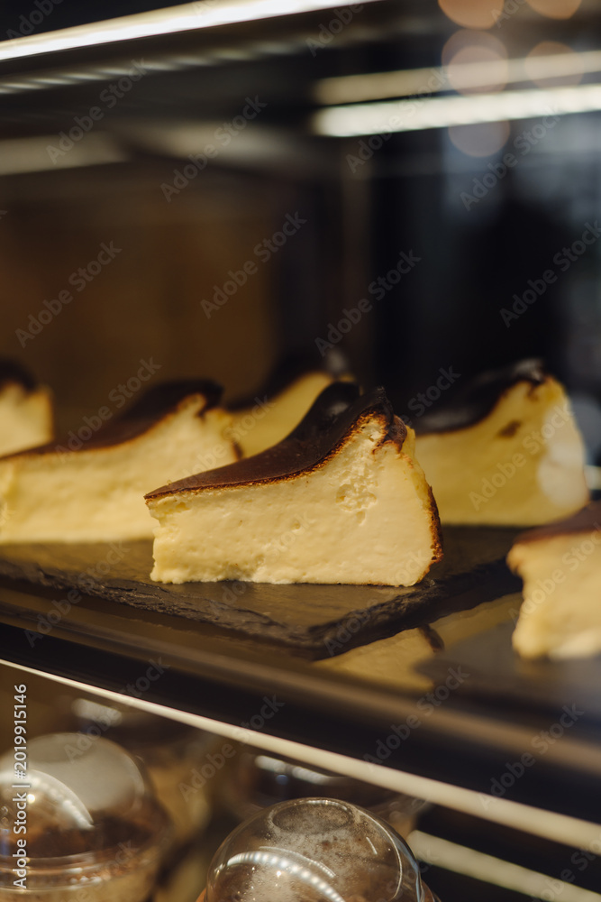 Fototapeta premium Slices of San Sebastian burnt cheesecake on a dark display shelf in a cafe. Creamy Basque dessert with a caramelized top, perfect for restaurant menus and food photography.
