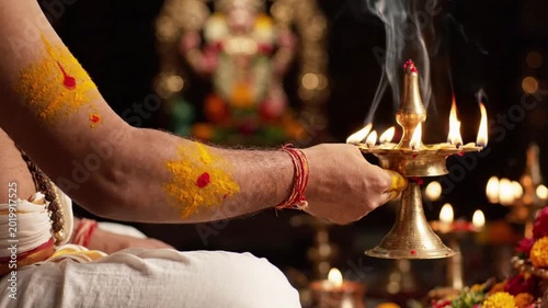 Hindu ritual aarti ceremony, hand holding a brass oil lamp with multiple wicks burning, smoke rising, traditional religious offering with yellow and red powder on arm, bokeh background