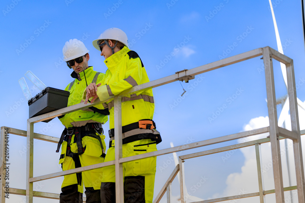 Fototapeta premium Professional wind turbine technicians in high-visibility safety gear using a laptop to monitor renewable energy operations on an elevated platform.
