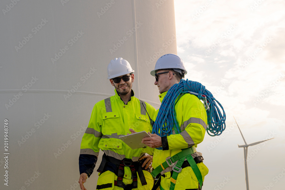 Fototapeta premium Two engineers in high visibility gear stand near a wind turbine, reviewing a clipboard and preparing for maintenance work. The image highlights teamwork, safety, and renewable energy sector.