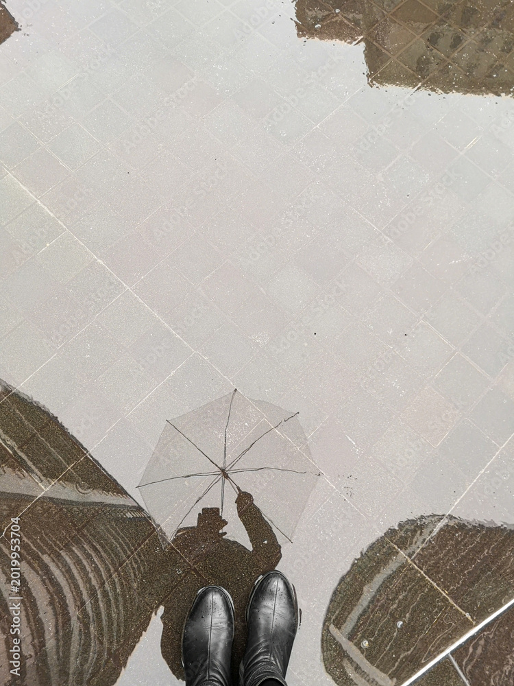 Obraz premium Reflection of person with umbrella in rain puddle outdoors. Wet city street, rainy weather, urban lifestyle, moody atmosphere.