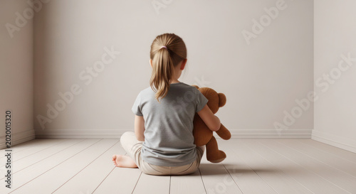 Young girl sitting on wooden floor holding teddy bear, facing empty room with light-colored walls and natural light streaming in. Lonely child feeling isolated, solitude, mental health, sadness.