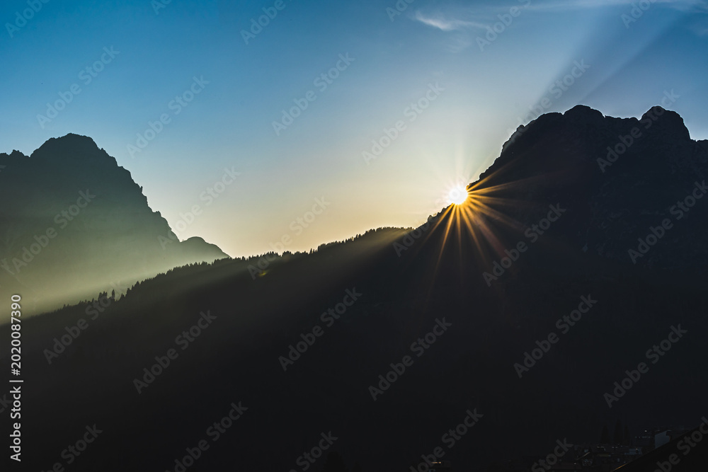 Obraz premium sole che tramonta dietro alle montagne, sagome scure delle catene montuose che vengono messe in risalto dalla luce del sole in un cielo sereno