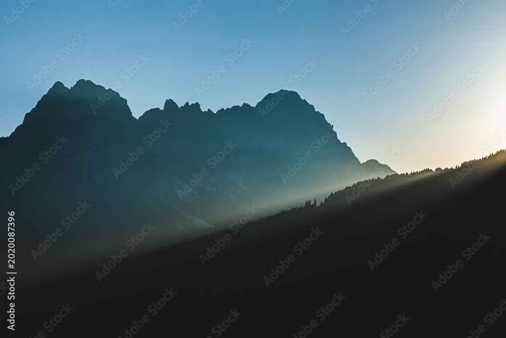 Obraz premium sole che tramonta dietro alle montagne, sagome scure delle catene montuose che vengono messe in risalto dalla luce del sole in un cielo sereno