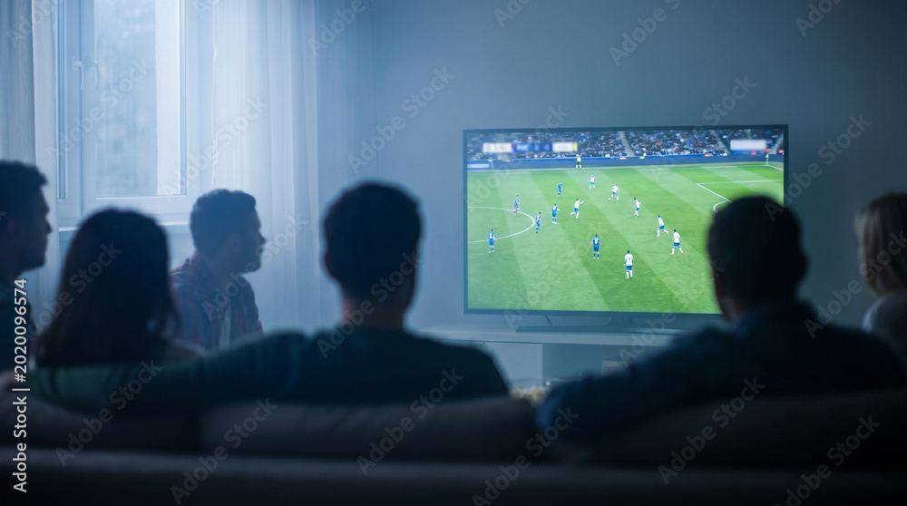 Fototapeta premium Group of friends watching soccer match on television in living room with soft lighting. Concept of global sports celebration, international football competition