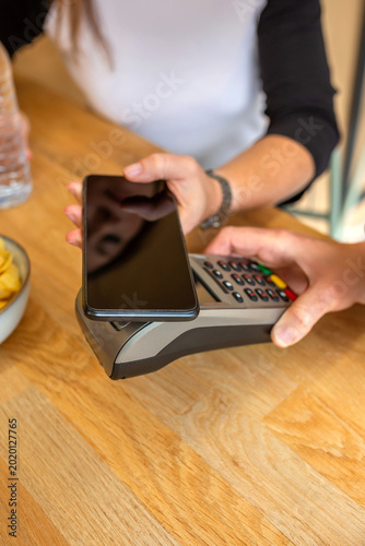 Anonymous woman is making a contactless payment using a smartphone over a point of sale pos terminal on a wooden counter, highlighting modern digital transactions and convenience