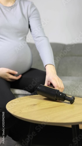 Vertical footage of pregnant woman using a percussion massage gun on her belly while sitting on a sofa.
