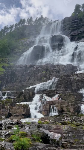 Panning video of Tvindefossen waterfall in Norway, with powerful white water cascading down layered rock formations surrounded by green vegetation under a dramatic cloudy sky