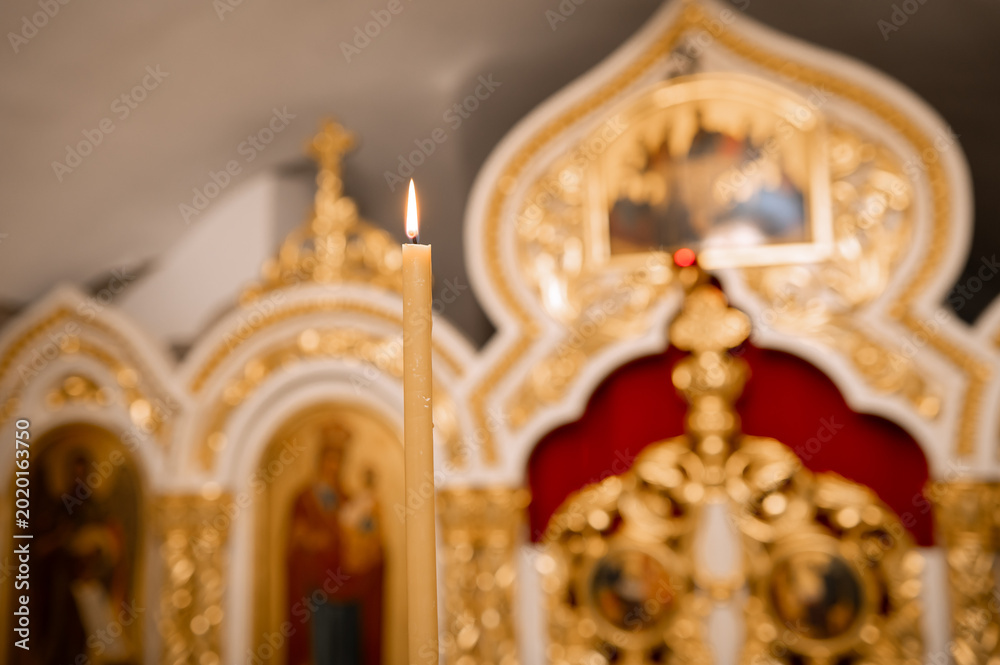 Fototapeta premium Burning wax candle close up with blurred golden iconostasis in Orthodox church interior