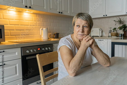 An elderly woman sits at the kitchen table in the morning  deep in thought with a serious expression on her face and her hands folded  looking away and deep in thought