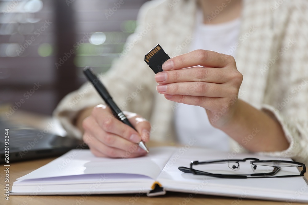 Fototapeta premium Woman with SD memory card taking notes at wooden table indoors, closeup