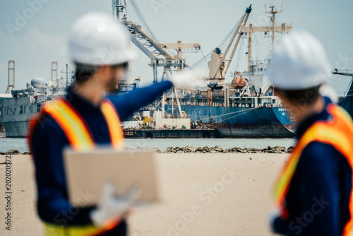 Logistics personnel in safety gear point toward a docking cargo vessel to direct maritime transport operations at a large commercial shipping harbor