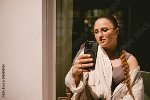 Smiling young woman with long braid and stylish glasses taking selfie on smartphone outdoors, happy female model posing for mobile photo or recording video blog in sunny European city street