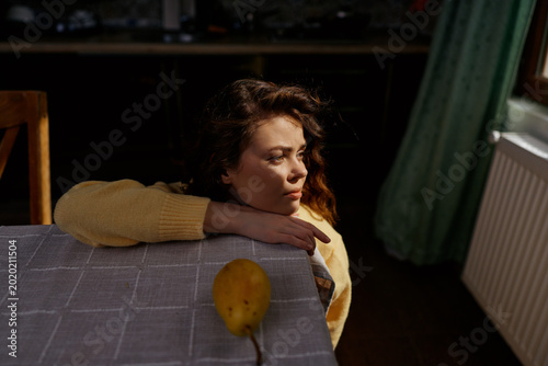 A contemplative young woman in a yellow sweater rests her head on a table beside a mango in a softly lit indoor scene