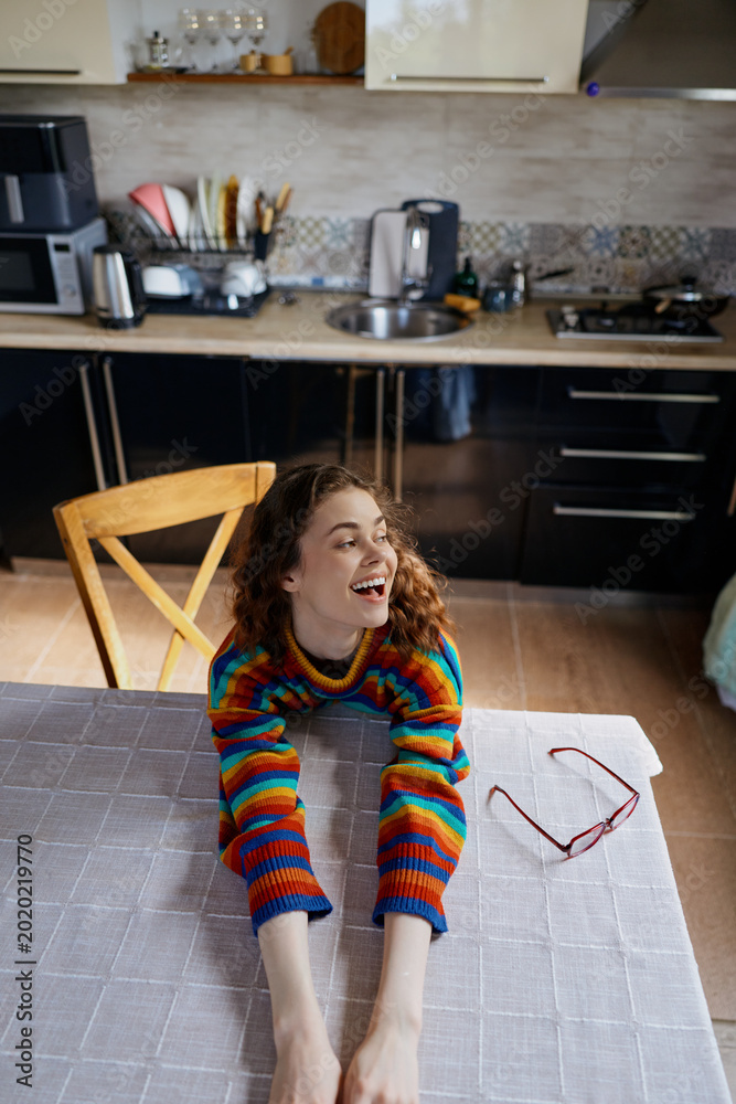 Fototapeta premium A young woman with curly hair wearing a colorful striped sweater smiles across a bright kitchen table, enjoying a casual home moment as sunlight streams in and sunglasses rest beside a laptop.