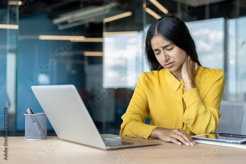 Tired young Indian woman sitting at the desk in the office, suffering from neck pain, getting a massage