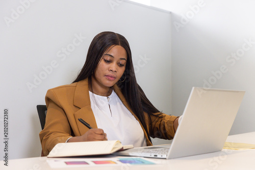 Midadult African American woman in tan blazer writing with pen and checking laptop swatch at desk