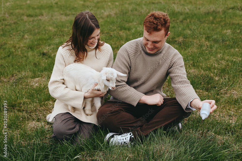 Obraz premium A couple sit on the grass outdoors, cuddling a white goat kid while a bottle feeds in a serene rural moment. A gentle, affectionate scene highlighting lifestyle, animal care, and family bonding in