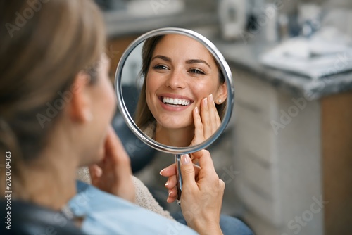 Happy woman admire smile in small mirror; dental patient satisfied with teeth whitening treatment