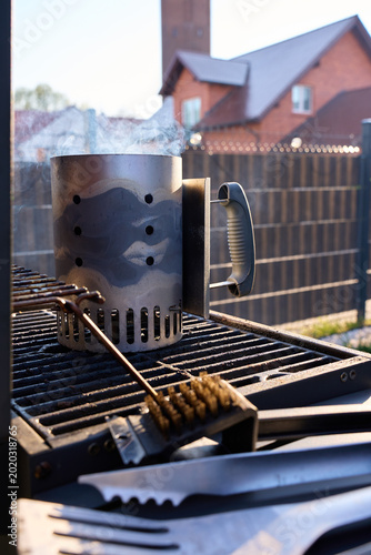 Fotografie Smoking charcoal chimney starter stands on grill grate with barbecue brush and f