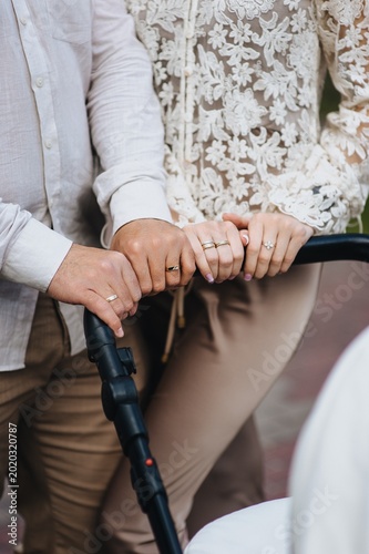 A close-up photograph of the hands of a young man, father, and mother holding onto a stroller with a newborn baby on a walk.