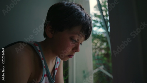 фотографія Young child sitting by window with downward gaze, pensive expression and quiet s