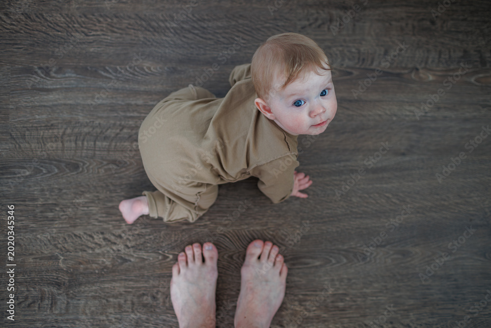 Naklejka premium Infant on all fours on a dark wooden floor looking up at a person standing nearby