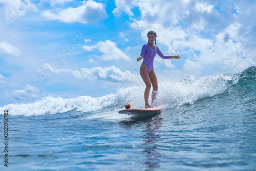Young Woman Surfing On Blue Wave, Balanced On Surfboard With Action Camera Mounted, Sunlit Sea And Cresting