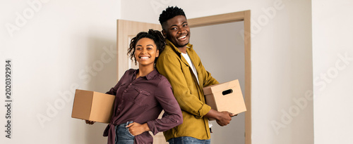 A couple carries brown boxes while smiling in a room. They stand back to back in a newly moved-in space. The sunlight shines through the open door behind them  showing a fresh start.