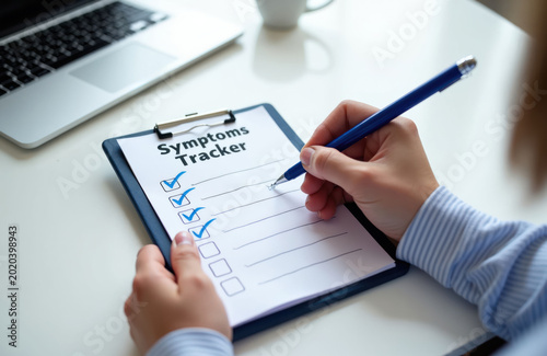 Person fills out symptoms tracker checklist with pen on clipboard. Hand marks items on health form. Laptop and coffee cup nearby. Doctor consults patient data on desk.