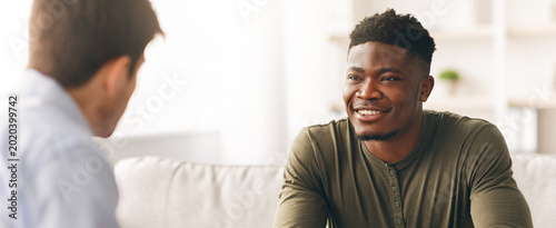 Two men sit across from each other in a bright room. They engage in conversation  exchanging smiles and nods. The space has soft furniture and warm light  contributing to a friendly atmosphere.