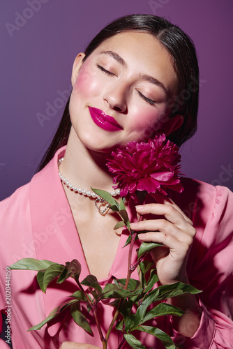 Young woman with closed eyes and bright pink lipstick holds dark pink peony flower close  wearing pink blazer and pearl necklace. Fashion and beauty creative portrait.