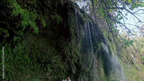 Close-up view of water flowing over mossy rocks in a lush forest, showing a peaceful waterfall scene with green vegetation, wet cliffs and natural daylight for travel and nature footage.