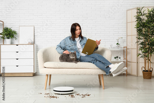 Tableau sur toile Young woman with cute cat and robot vacuum cleaner reading magazine at home