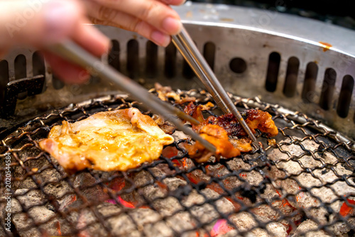 Close up of meat grilling on a charcoal barbecue grill with hands using metal chopsticks to turn the food. Traditional yakiniku or Korean BBQ dining experience with glowing embers and smoke.