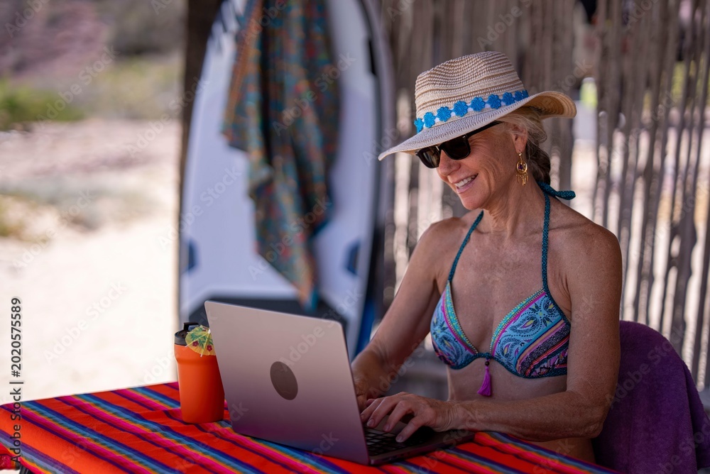 Fototapeta premium Mature woman sits at a table on a tropical beach, using a computer while smiling, enjoying her vacation and the warm weather