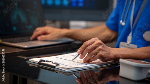Foto Close-up of healthcare worker in blue scrubs writing on clipboard beside laptop in hospital, medical data on monitors, clinical workspace, focused documentation and patient care concept