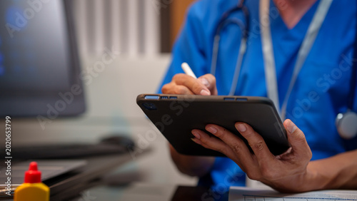 Vászonkép Close-up of heaalthcare worker in blue scrubs writing on a digital tablet with stylus at desk, stethoscope visible, computer monitor blurred in background, modern clinic office