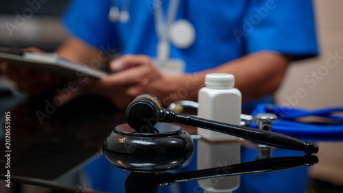 Fotografie Medical malpractice legal concept with judge gavel on desk, prescription bottle and stethoscope, doctor in scrubs holding clipboard in background