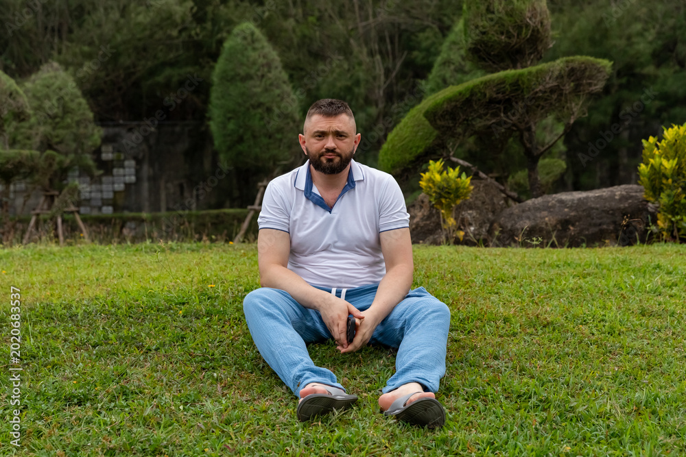 Obraz premium Man with beard sitting on green grass in a park, surrounded by trees and shrubs, wearing casual clothing and sandals, enjoying a relaxed outdoor moment