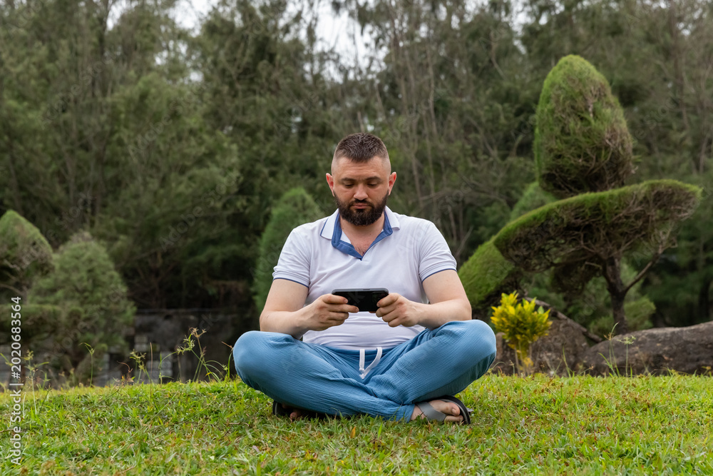 Obraz premium Man sitting cross-legged on green grass, focused on a smartphone in his hands, surrounded by a landscaped garden featuring neatly trimmed shrubs and trees in the background